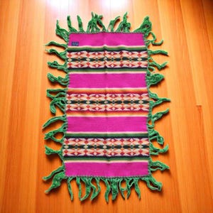 May include: A pink and green woven blanket with fringe. The blanket has a geometric pattern in shades of orange, yellow, green, and black.