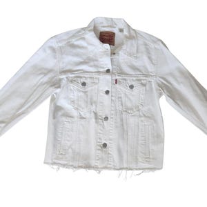 Puede incluir: Chaqueta vaquera blanca con un diseño clásico. Cuenta con cierre de botones, dos bolsillos en el pecho y dos bolsillos laterales. La chaqueta tiene un dobladillo deshilachado y una etiqueta roja de Levi's. Una prenda versátil.