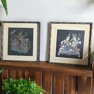 Könnte beinhalten: Zwei gerahmte Kunstwerke im asiatischen Stil. Die Kunstwerke zeigen farbenfrohe Figuren auf dunklem Hintergrund. Die Rahmen haben ein bambusähnliches Design. Die Drucke werden auf einem Holzregal ausgestellt.