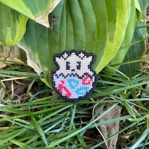 May include: A small, cross-stitched patch featuring a cartoon character with a white body, black outline, and a blue and red design. The patch is set against a backdrop of green leaves and grass.