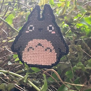 May include: A black and white cross-stitch of a cartoon character with large eyes and a small mouth. The character is sitting on a green leafy background.