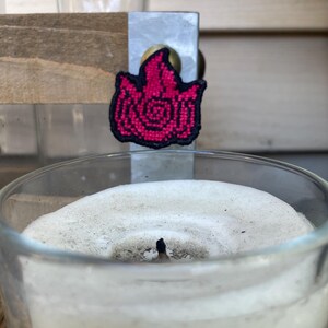 May include: A close-up of a white candle with a black wick in a clear glass jar. A small pink and black embroidered patch of a flame is attached to the jar.