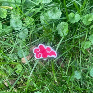 May include: A red cloud-shaped patch with white trim, resting in a bed of green grass and leaves. The patch has a pixelated design, with a series of white shapes inside the red cloud. The grass is a mix of long blades and small, round leaves.