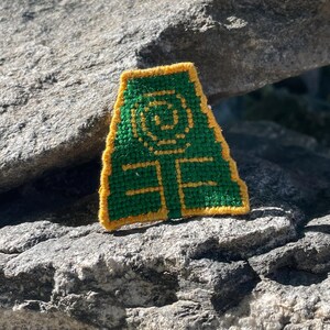 May include: Green and yellow embroidered patch with a stylized symbol of a triangle with a circle inside. The symbol is surrounded by a yellow border.