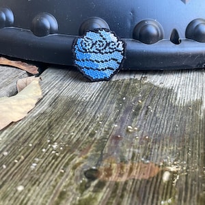 May include: A blue and black embroidered patch with a stylized image of a planet with rings. The planet is surrounded by a white border.