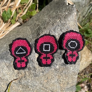 May include: Three red and black embroidered patches featuring the Squid Game guards. Each patch has a different shape on the mask: a triangle, a square, and a circle. The patches are displayed on a gray rock.