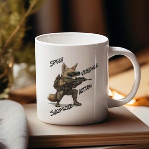May include: White ceramic mug featuring a cartoon fox in military gear, holding a rifle. The mug has the words "SPEED", "VIOLENCE", "ACTION", and "SURPRISE" in black script. The mug is sitting on a stack of books.