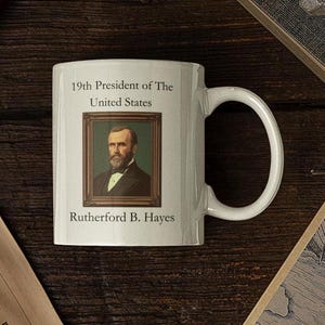 Op de afbeelding: Een witte keramische mok met een zwart-wit portret van Rutherford B. Hayes, de 19e president van de Verenigde Staten. De tekst "19th President of The United States" en "Rutherford B. Hayes" staat op de mok gedrukt.