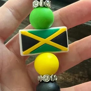 May include: A keychain with a Jamaican flag design. It features black and yellow beads, a rectangular charm with the flag's colours, and a silver-coloured clasp.