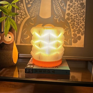 May include: A decorative lamp with a ribbed, cylindrical shade, emitting a warm glow. The lamp sits on a stack of books with an orange base. A felt owl and green plant are visible in the background. The lamp is a stylish home decor item.