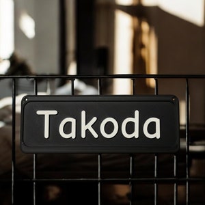 May include: A black rectangular sign with the name "Takoda" in white, mounted on a black metal cage. The sign has rounded corners and is centered in the frame. The background is blurred.