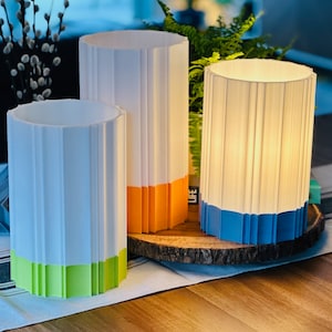 May include: Three cylindrical lamps with a ribbed design. The lamps are white with colored bases in green, orange, and blue. The lamps are arranged on a wooden slice, with greenery in the background. The lamps are lit from within.