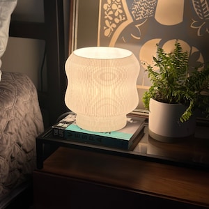 May include: A white ceramic table lamp with a ribbed design, turned on and illuminating a bedside table with a book titled "The Life" and a potted fern plant.