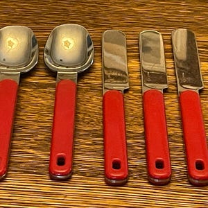 May include: A set of five stainless steel serving utensils with bright red handles. The set includes two ice cream scoops and three butter knives. The handles have a hole at the end for hanging.