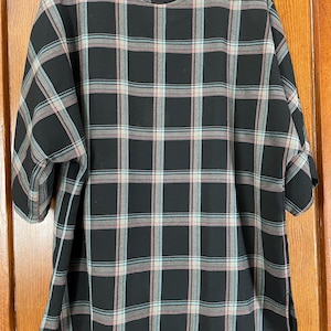 May include: A black plaid top with short sleeves, featuring a pattern of intersecting lines in light blue, pink, and white. The top has a round neckline and is fastened with two buttons on the shoulder. The fabric appears to be lightweight.