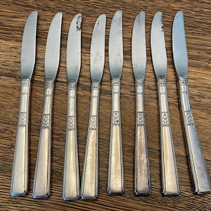 May include: Eight silver butter knives with rounded blades and patterned handles. The knives are displayed on a wooden surface. Each handle features a geometric design, adding a touch of elegance to the cutlery set, suitable for dining.