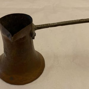 May include: A copper-colored, vintage Turkish coffee pot, also known as a cezve, with a long, ornate handle. The pot has a wide base that tapers to a narrow neck and a flared rim. The handle features a decorative floral design.