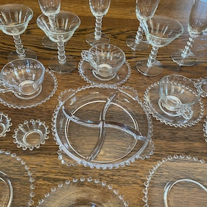 May include: A collection of clear glass tableware, including stemmed glasses, teacups with saucers, and plates. The glasses have a distinctive beaded stem design. The plates and saucers feature a beaded edge. A divided serving dish is also present.