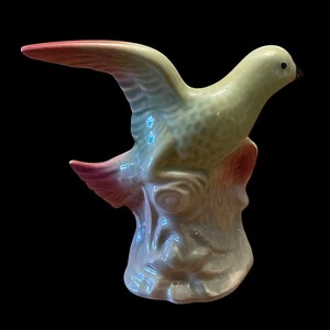 May include: A ceramic bird figurine in flight, perched on a tree stump. The bird features a gradient of colors, with pink and red wing tips, transitioning to light green and blue. The base is a light gray color.