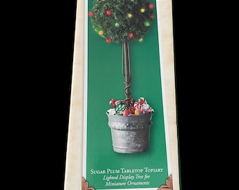 May include: A box featuring a Sugar Plum tabletop topiary. The display tree has a green, round top with red, yellow, and green lights. The tree sits in a silver bucket filled with miniature ornaments. Text on the box reads "Lighted Display Tree for Miniature Ornaments."