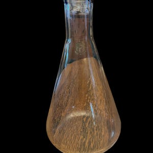 May include: A clear glass Erlenmeyer flask with a wide, rounded base and a narrow neck. The flask has a textured interior and a small emblem near the top. The flask is empty and sits against a black background.