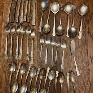 May include: An assortment of vintage silverware, including forks, spoons, knives, and serving spoons, arranged on a wooden surface. The silverware has a tarnished, antique appearance, with ornate handle detailing. The collection includes various sizes and styles of utensils.