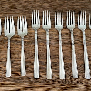 May include: A collection of nine silver forks with three tines each, arranged on a wooden surface. The forks have decorative patterns on the handles and are evenly spaced. The tines are slightly curved, and the overall design is classic.