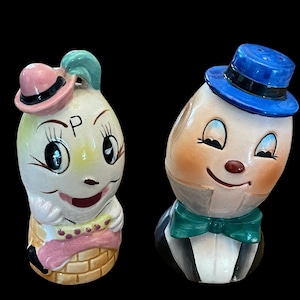 May include: A pair of vintage ceramic salt and pepper shakers. One shaker is a white onion-shaped figure wearing a pink hat and a basket-like base. The other is an egg-shaped figure in a blue hat, bow tie, and tuxedo.