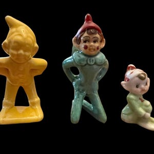 May include: Three ceramic figurines of gnomes in various poses. The first gnome is yellow with arms akimbo. The second gnome is teal with a red hat. The third gnome is light green and seated. All have painted faces.