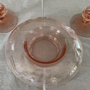 May include: A set of three vintage pink glass candle holders with etched floral designs. The candle holders are in the shape of a bowl with a raised center for the candle.