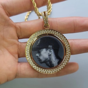 May include: A gold-toned circular pendant necklace featuring a rotating centre disc with a black and white engraved portrait of a person wearing a baseball cap. The outer frame and the bail are encrusted with small clear stones on a rope chain.