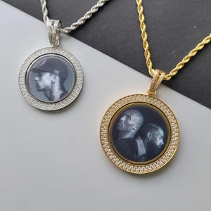 May include: Two circular pendants on rope chains, one silver-toned and one gold-toned. Each pendant features a border of small clear stones surrounding a central black and white photographic portrait of individuals, suspended from a matching metal chain.