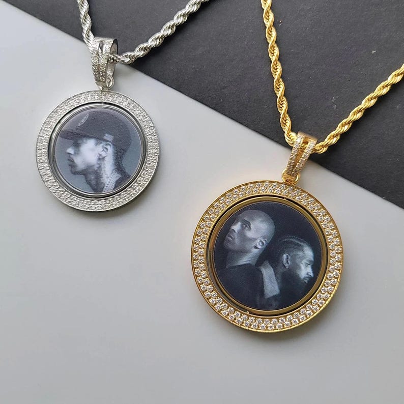 May include: Two circular pendants on rope chains, one silver-toned and one gold-toned. Each pendant features a border of small clear stones surrounding a central black and white photographic portrait of individuals, suspended from a matching metal chain.