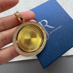 May include: A circular gold-tone pendant featuring a central smooth gold disc surrounded by a border of small clear stones. The pendant has a decorative bail encrusted with clear stones and is positioned above a blue box labelled JOY & RACHEL.