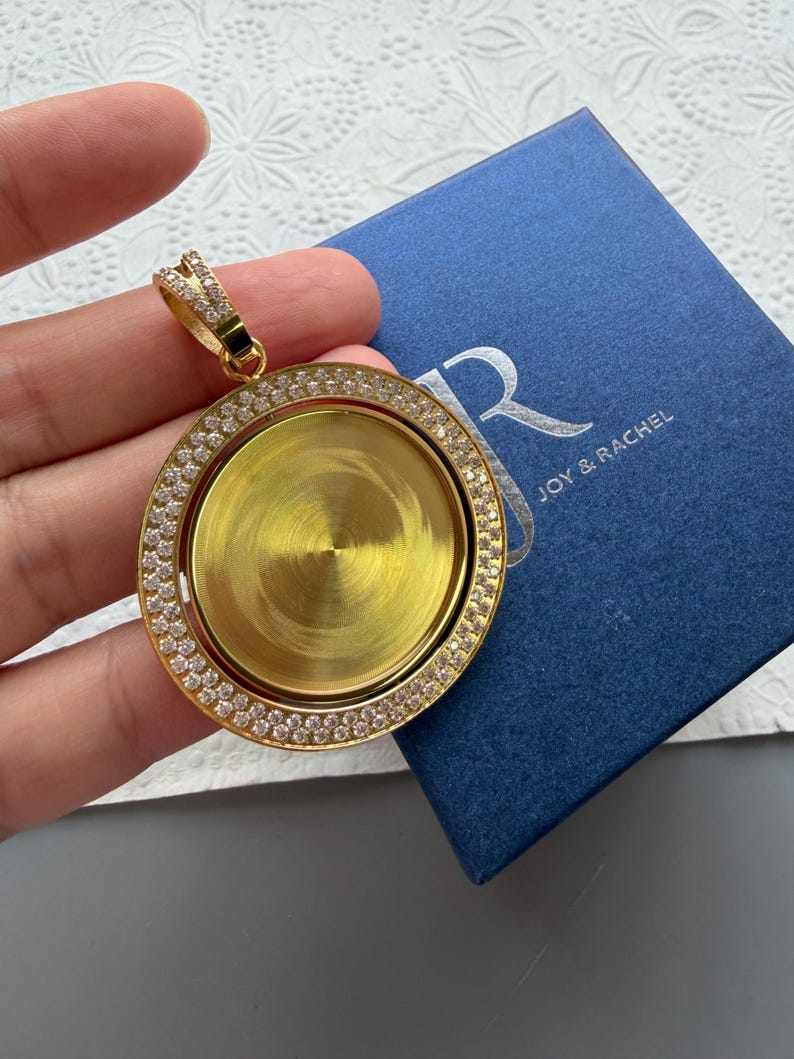 May include: A circular gold-tone pendant featuring a central smooth gold disc surrounded by a border of small clear stones. The pendant has a decorative bail encrusted with clear stones and is positioned above a blue box labelled JOY & RACHEL.