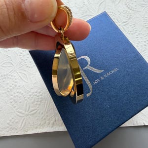 May include: A gold-toned pendant featuring a circular frame with a translucent centre, suspended from a small bail with embedded stones. The pendant is positioned above a dark blue square box with the silver text JOY & RACHEL printed on the lid.