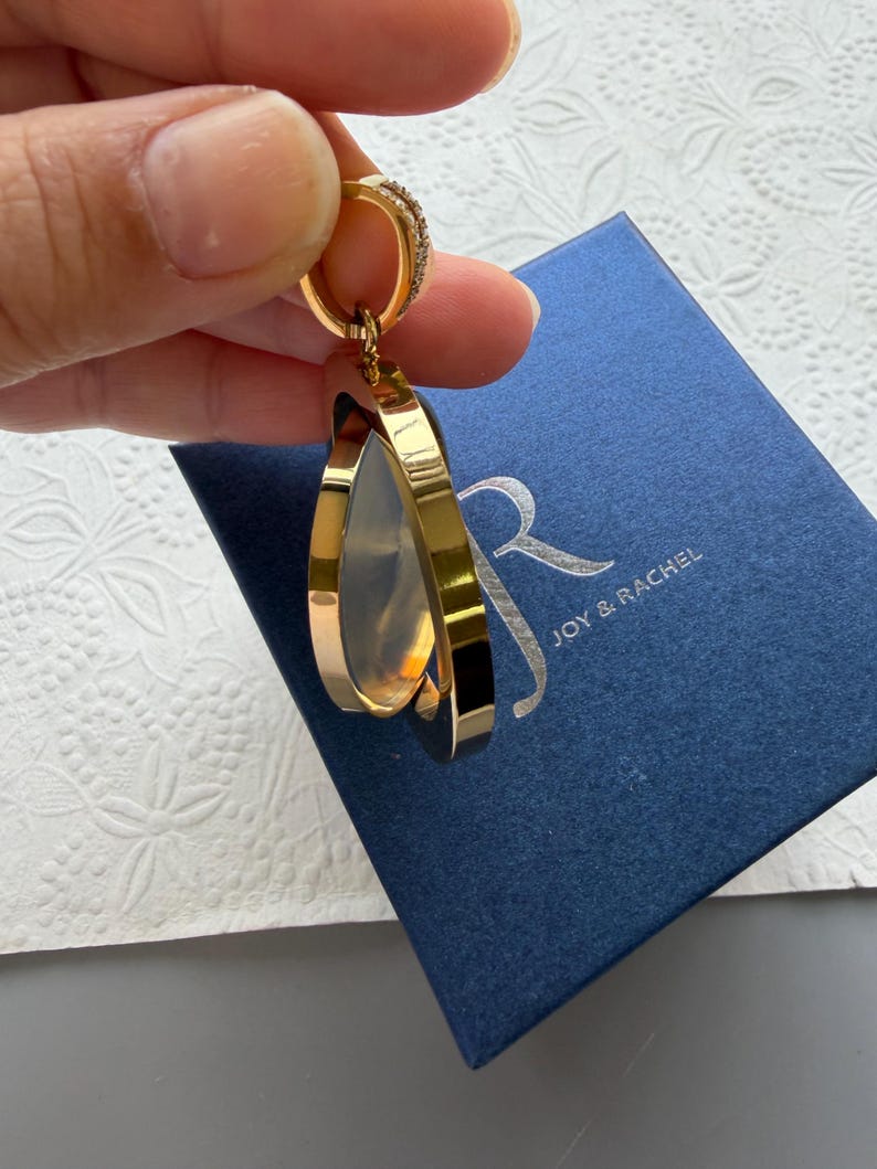 May include: A gold-toned pendant featuring a circular frame with a translucent centre, suspended from a small bail with embedded stones. The pendant is positioned above a dark blue square box with the silver text JOY & RACHEL printed on the lid.