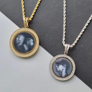 May include: Two circular pendant necklaces with rope chains. One is gold-toned with a black and white portrait of two individuals. The other is silver-toned with a black and white portrait of one individual wearing a cap. Both feature crystal-set borders.
