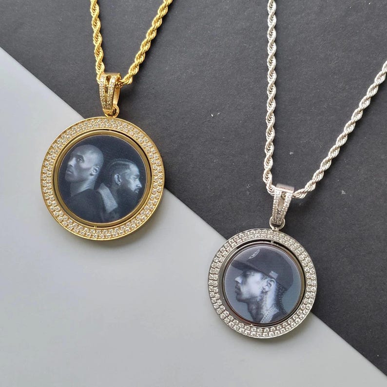 May include: Two circular pendant necklaces with rope chains. One is gold-toned with a black and white portrait of two individuals. The other is silver-toned with a black and white portrait of one individual wearing a cap. Both feature crystal-set borders.
