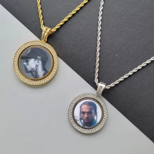 May include: Two circular pendants on rope chains. The gold-tone pendant features a black and white portrait of a person in a baseball cap. The silver-tone pendant features a colour portrait of a person with braids, both framed by clear stones.