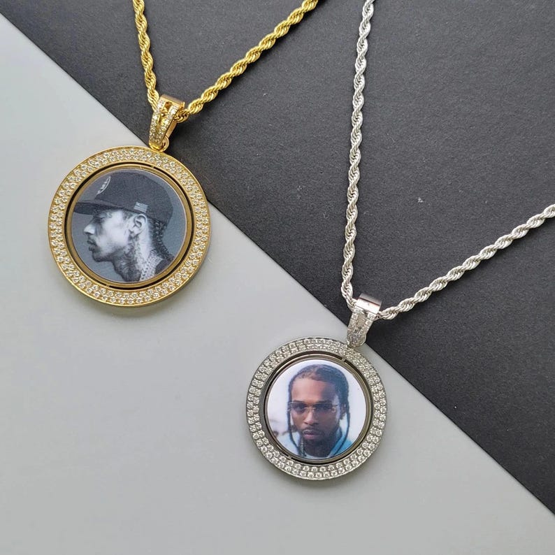 May include: Two circular pendants on rope chains. The gold-tone pendant features a black and white portrait of a person in a baseball cap. The silver-tone pendant features a colour portrait of a person with braids, both framed by clear stones.