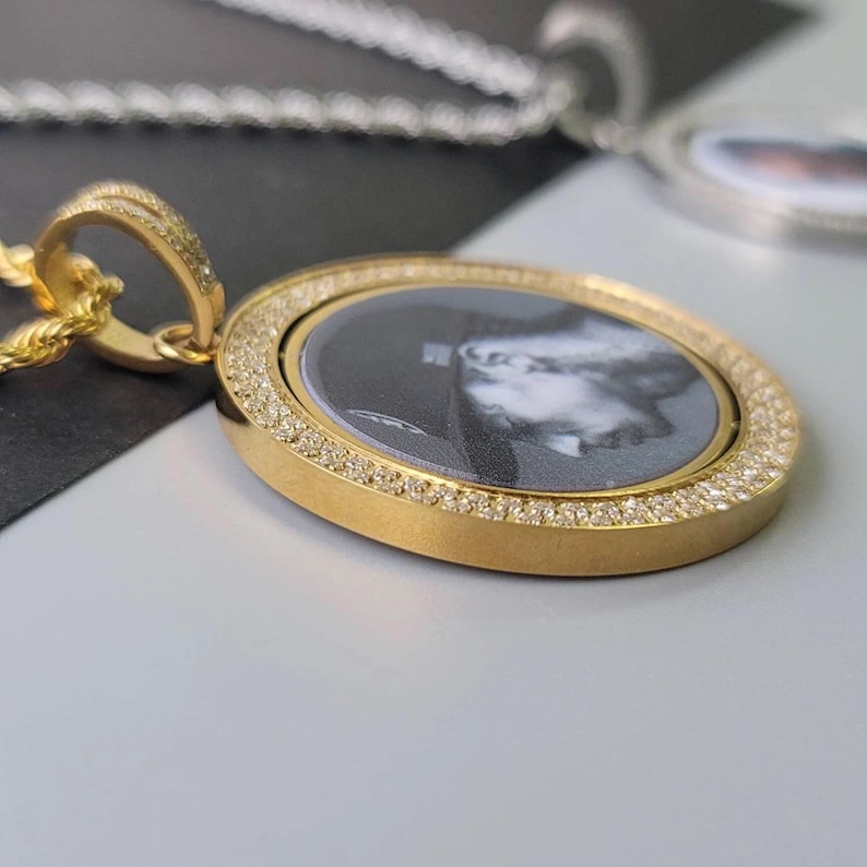 May include: A gold-toned circular pendant necklace featuring a central black and white portrait insert. The pendant is framed by a border of small, clear stones and is attached to a gold-toned rope chain. A silver-toned chain is visible in the background.