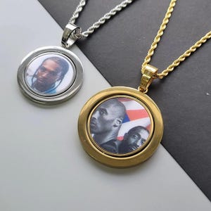May include: Two circular pendant necklaces with rope chains. The silver-tone pendant features a portrait of a person. The gold-tone pendant displays a black and white graphic of two individuals against a background with red, white, and blue stripes.
