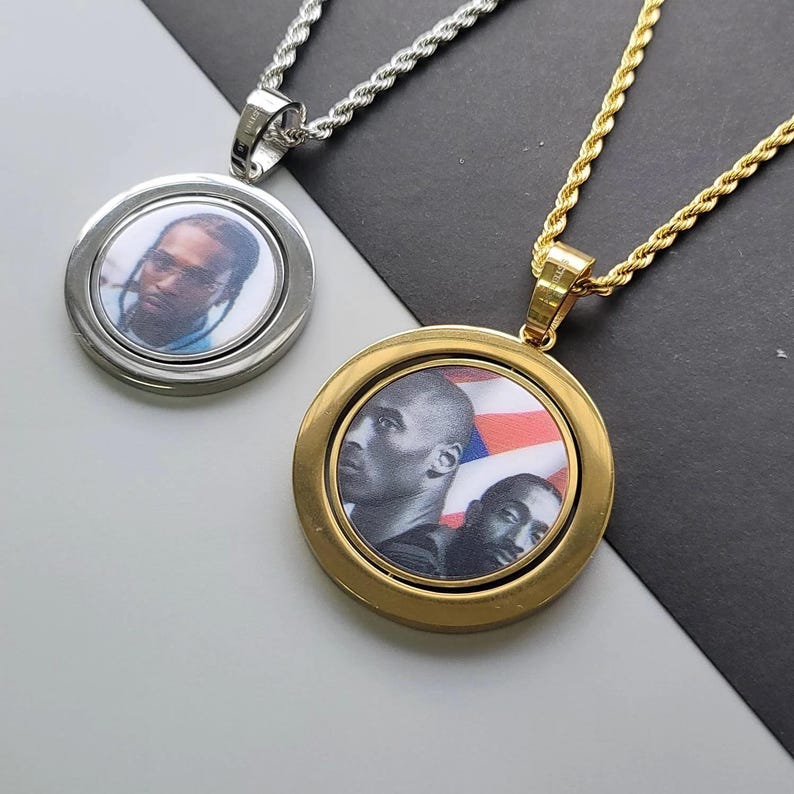 May include: Two circular pendant necklaces with rope chains. The silver-tone pendant features a portrait of a person. The gold-tone pendant displays a black and white graphic of two individuals against a background with red, white, and blue stripes.