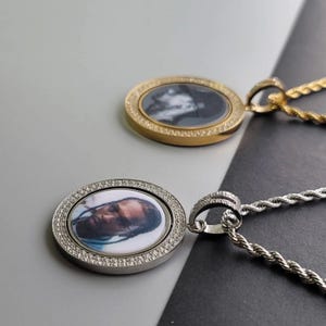 May include: Two circular pendants on rope chains, one gold-toned and one silver-toned. Each pendant features a border of small clear stones surrounding a central circular portrait of a person. The silver pendant displays a clear colour photograph.