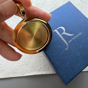 May include: A circular gold-toned pendant with a brushed metallic centre and a polished outer rim, featuring a small bail at the top. The pendant is positioned next to a square blue gift box with the silver text Joy & Rachel printed on the lid.