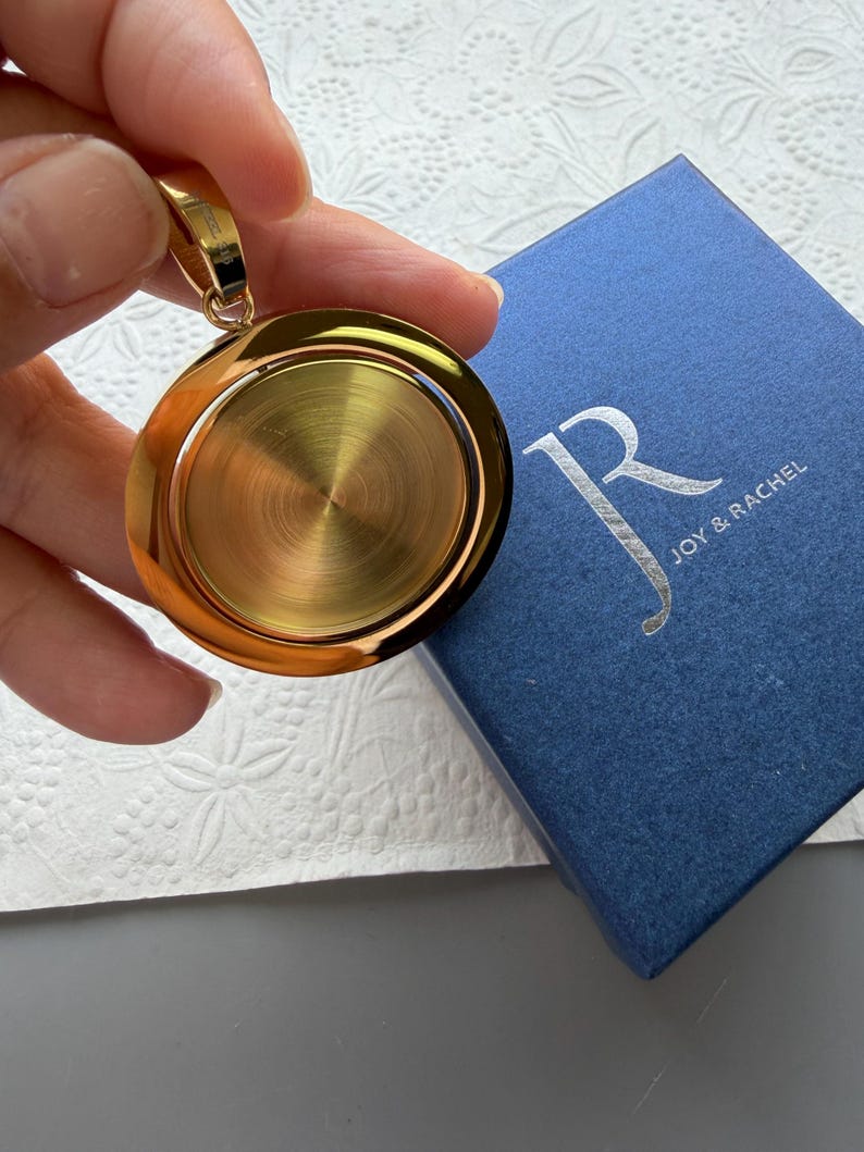 May include: A circular gold-toned pendant with a brushed metallic centre and a polished outer rim, featuring a small bail at the top. The pendant is positioned next to a square blue gift box with the silver text Joy & Rachel printed on the lid.