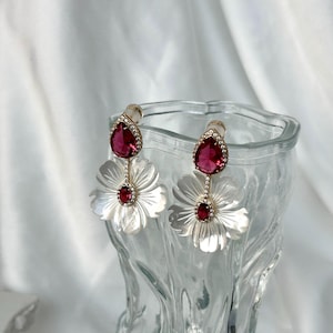 May include: A pair of white flower-shaped earrings with a red teardrop-shaped gemstone in the center of each flower. The earrings are made of mother of pearl and have a gold-tone finish.