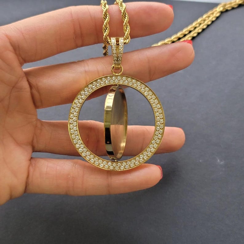 May include: A gold-toned necklace featuring a circular pendant encrusted with small clear stones. The pendant contains a central rotating disc element suspended from a thick rope chain. The entire piece is finished in a polished gold-tone metal.