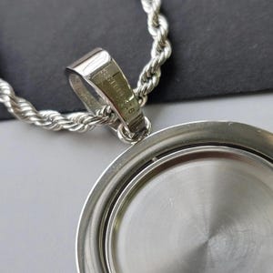 May include: A silver-toned metal pendant with a circular, recessed centre and a thick, polished bail. The bail is engraved with the text S.STEEL 316. The pendant is attached to a silver-toned rope chain with a twisted, textured link design.